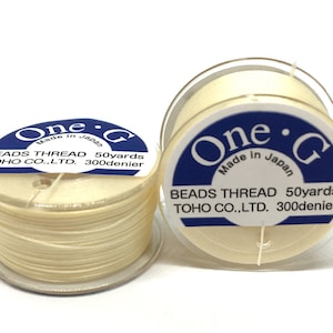 May include: Two spools of cream-coloured bead thread are shown against a white backdrop. Each spool has a clear plastic base and a blue and white label that reads "One G Made in Japan BEADS THREAD 50 yards TOHO CO., LTD. 300 denier."