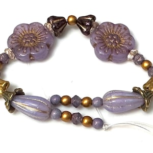 May include: A delicate bracelet featuring lavender flower and teardrop-shaped beads, accented with gold and dark purple beads. The bracelet is strung on a clear cord, showcasing a vintage-inspired design.