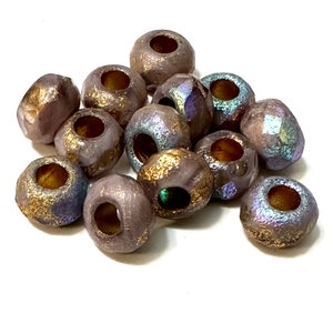 May include: A selection of faceted, barrel-shaped beads. The beads feature a lavender-grey base colour with gold and iridescent accents. Each bead has a large hole through the centre, suitable for stringing. These beads are likely for jewellery making.