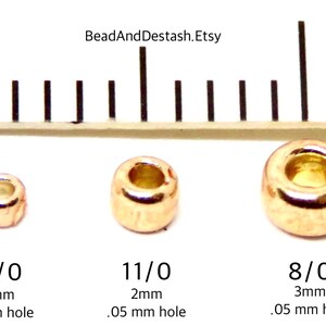 Op de afbeelding: Drie goudkleurige metalen kralen van verschillende maten worden naast een liniaal getoond. De kralen zijn gemarkeerd met 15/0 (1,3 mm), 11/0 (2 mm) en 8/0 (3 mm), met een gatgrootte van 0,05 mm. De Etsy shopnaam BeadAndDestash.Etsy is zichtbaar.