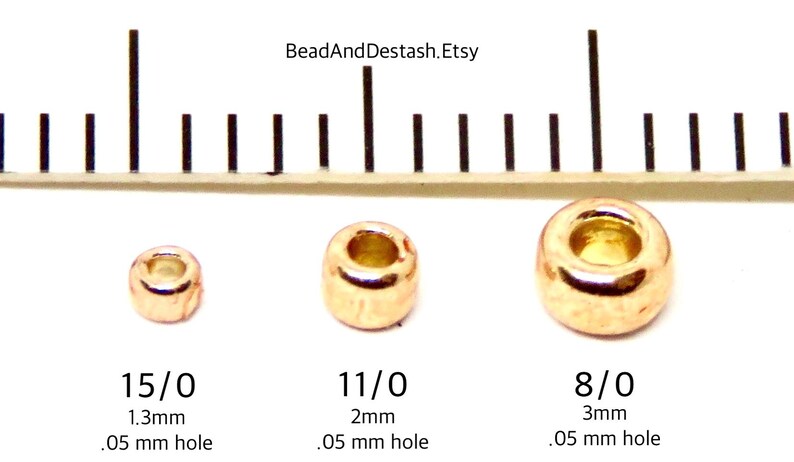 Op de afbeelding: Drie goudkleurige metalen kralen van verschillende maten worden naast een liniaal getoond. De kralen zijn gemarkeerd met 15/0 (1,3 mm), 11/0 (2 mm) en 8/0 (3 mm), met een gatgrootte van 0,05 mm. De Etsy shopnaam BeadAndDestash.Etsy is zichtbaar.