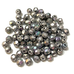 May include: A collection of faceted, metallic grey beads with iridescent highlights. The beads have multiple sides and small holes for stringing. The beads are scattered on a white surface.