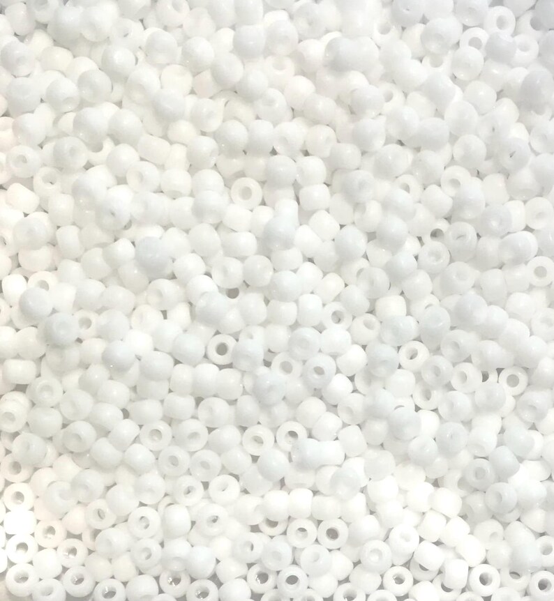 Op de afbeelding: Een close-up van een stapel kleine, ondoorzichtige, witte zaadkralen. De kralen zijn uniform van grootte en vorm, met een klein gaatje in het midden. Ze zijn verspreid, waardoor een textuur ontstaat.