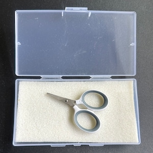 May include: Small, silver scissors with white and grey handles, inside an open clear plastic case. The case reveals the scissors on a bed of light-coloured foam. The scissors are designed for precision cutting, ideal for crafts.