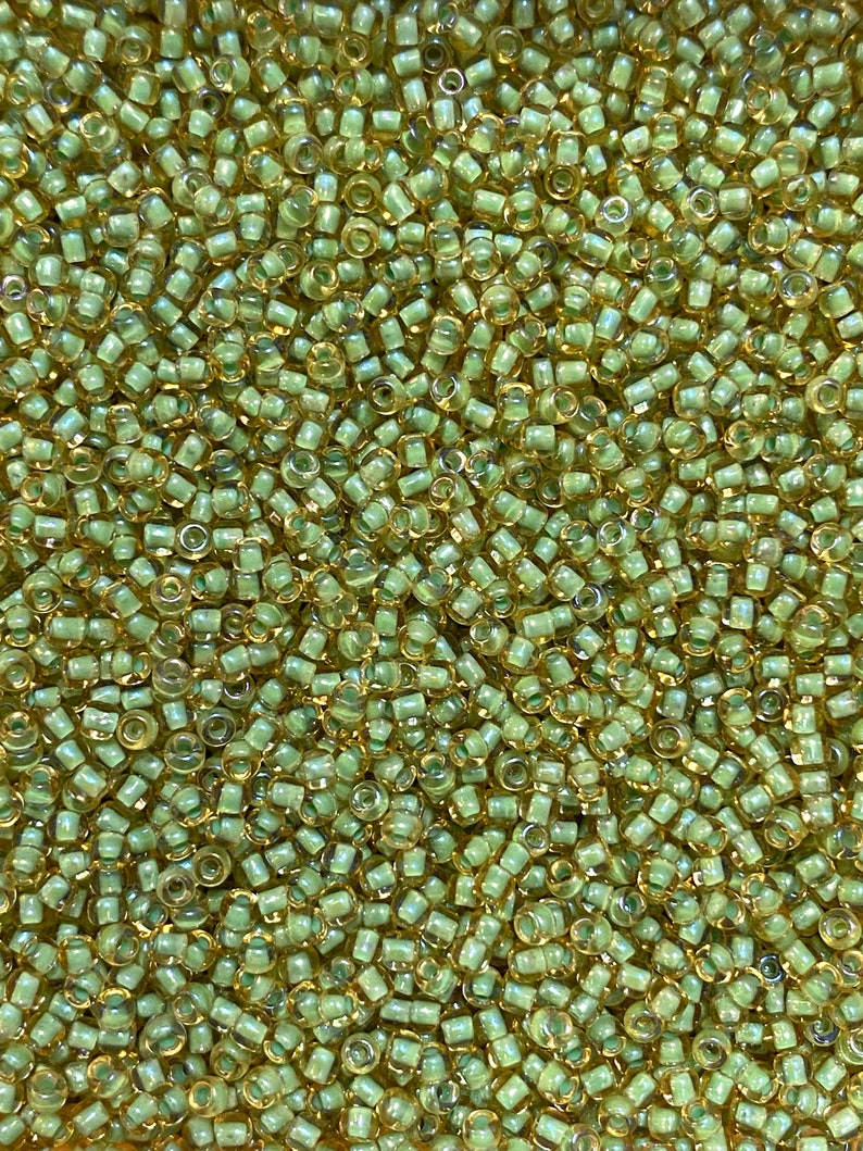 K&ouml;nnte beinhalten: Nahaufnahme eines Haufens kleiner, zylindrischer Perlen in Hellgr&uuml;n und Gold. Die Perlen sind dicht gepackt und bilden eine strukturierte Oberfl&auml;che. Die Perlen werden wahrscheinlich f&uuml;r die Schmuckherstellung oder andere Bastelprojekte verwendet.