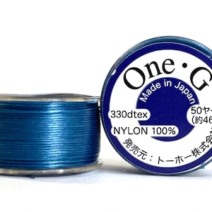 Peut inclure: Une bobine de fil de nylon bleu avec le texte "One G Made in Japan". L'étiquette indique également 330dtex, environ 46 mètres et "NYLON 100%".