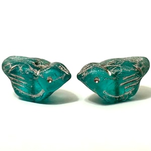 May include: Two teal bird-shaped glass beads with silver detailing. The beads have a textured surface and small holes for threading. The bird's wings and head are detailed with silver lines. These beads are suitable for jewellery making.