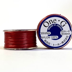 Peut inclure: Bobine de fil rouge en nylon. L'étiquette indique "One G Made in Japan", "330dtex" et "50 yards (environ 46m)". Le fil est composé à 100% de nylon.