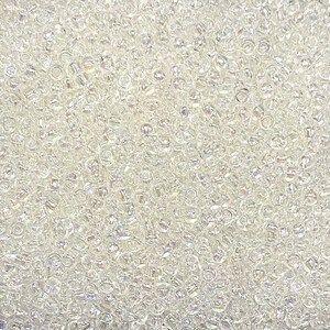 May include: Close-up of a pile of small, clear glass seed beads. The beads are cylindrical with a hole running through the centre. The beads are scattered and reflect light, creating a sparkling effect.