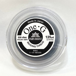May include: A spool of dark gray nylon beading thread, labeled "One G" and "TOHO BEADS." The label also indicates 125 yards (114 m) of thread, made in Japan. The thread is 330 dtex, and the spool is in a clear plastic container.