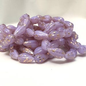 Peut inclure: Un tas de perles en forme de goutte, de couleur lavande, avec des éclats dorés. Les perles ont une finition brillante et sont regroupées sur un fond blanc. Idéales pour la création de bijoux.