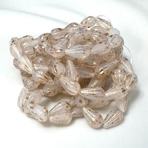 May include: A collection of clear, teardrop-shaped beads with gold flecks. The beads have vertical ridges and are strung together. The beads are a pale pink colour and are on a white background. Suitable for jewellery making.