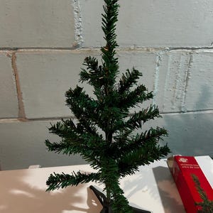 May include: A miniature artificial Christmas tree with dark green needles and a black plastic base. The tree is on a white surface, with a red box labelled "Christmas tree" in English and French in the background.