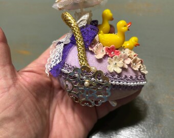 May include: A purple pincushion with a white lace trim and a gold cord handle. The pincushion is decorated with pink and white flowers and three yellow rubber duckies. The pincushion is sitting on a hand.