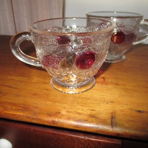 Puede incluir: Dos tazas de vidrio transparente con un patrón en relieve y acentos de fruta de vidrio rojo. Las tazas tienen un asa y una base redondeada.