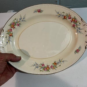 May include: Oval serving platter with a cream-colored surface and a gold rim. The platter is decorated with floral arrangements in pink, yellow, and blue. The edges have a decorative design.