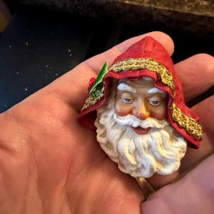 May include: A Santa Claus head figurine featuring a red hood trimmed with gold metallic braid and a small green holly leaf with a red berry accent. The figure has a white beard, moustache, and blue eyes, with a painted finish on the face.