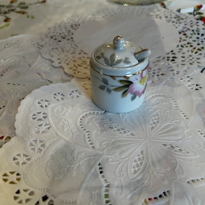 May include: A white porcelain sugar bowl with a floral design and a gold trim. The bowl has a lid and a small spoon. The bowl is sitting on a white doily.