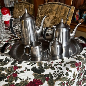 Könnte beinhalten: Ein Edelstahl-Teeservice auf einem silbernen Tablett. Das Set umfasst zwei Teekannen, einen Sahnekännchen und eine Zuckerdose. Das Tablett hat schwarze Griffe. Das Set steht auf einer Tischdecke mit einem Stechpalmen- und Beerenmuster.