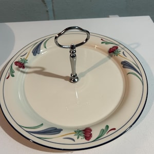 May include: A two-tiered serving plate with a silver handle. The off-white plates have a blue border and are decorated with red and blue floral patterns. The handle is a loop shape.
