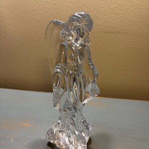 Op de afbeelding: Een helder glazen engelenbeeldje met gedetailleerde vleugels en vloeiende gewaden. De engel staat op een getextureerde basis, met licht dat weerkaatst op het glas. Het beeldje is een decoratief item.