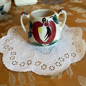 May include: A small white ceramic creamer with a red apple design. The creamer has two handles and is decorated with green leaves and a brown stem.