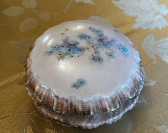 May include: A two-piece porcelain trinket box with a scalloped edge. The top piece is white with a floral design of blue forget-me-nots. The bottom piece is a darker shade of pink.