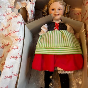 Könnte beinhalten: Eine Shirley Temple-Puppe in einer Box. Die Puppe hat blonde Locken und trägt eine weiße Bluse, eine schwarze Weste mit roter Schnürung, einen grün-weiß gestreiften Rock und einen roten Rock. Die Puppe trägt schwarze Strümpfe und braune Schuhe.