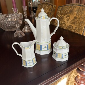 May include: A white porcelain tea set with a geometric design in yellow, blue, and green. The set includes a teapot, creamer, and sugar bowl. The teapot has a tall, slender spout and a matching lid. The creamer has a curved handle, and the sugar bowl has a lid.