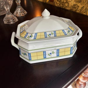 Puede incluir: Fuente blanca con tapa y estampado de cuadros amarillos y azules. La fuente tiene forma geométrica con un asa a cada lado. La tapa tiene un pequeño pomo cónico blanco.