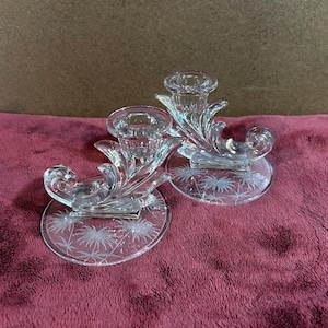 May include: Two clear glass candle holders with a floral design. Each holder has a circular base with an etched palm tree design. The candle holders have a decorative, swirling design and a cup to hold a candle.