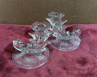 May include: A pair of clear glass candle holders with a floral design. Each holder has a circular base with an etched palm tree design. The candle holders have a decorative, swirling design and a cup to hold a candle.