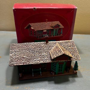 以下が含まれることがあります： 茶色の屋根と緑のトリムが付いた、ミニチュアの赤い駅。モデルには小さなベンチ、ミニチュアの木、前面に「Hays」の文字が含まれています。同様の画像が描かれた赤い箱が背景にあります。