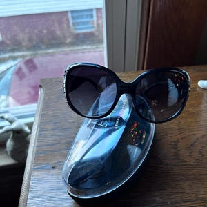 May include: Black oversized sunglasses with dark lenses rest on a clear case. The sunglasses have a rounded shape and decorative accents on the frames. The case has the words "ARMANI EXCHANGE" printed on it.