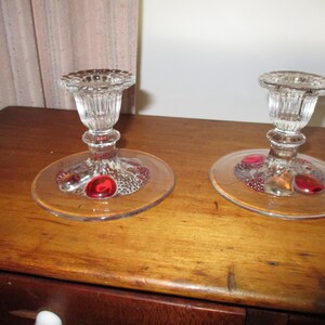 May include: Two clear glass candle holders with red and clear glass accents on the base. The candle holders are on a wooden surface.