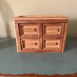 May include: A handcrafted wooden chest with four drawers. The chest is made of light and dark brown wood, with a rectangular shape and a slightly angled design. The drawers have simple, rectangular handles.