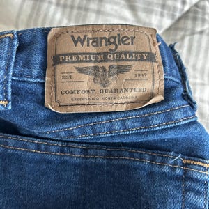 Pu&ograve; includere: Una toppa di denim blu con cuciture marroni e il testo "Wrangler Premium Quality Est. 1947 Comfort Guaranteed Greensboro, North Carolina"