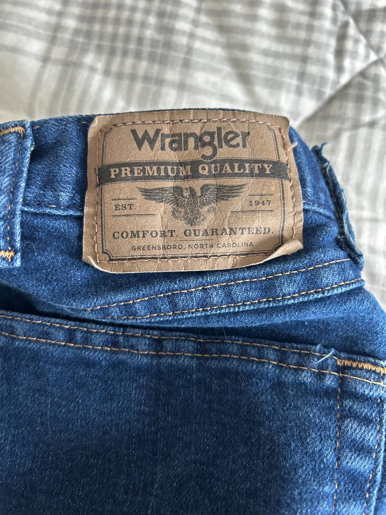 Pu&ograve; includere: Una toppa di denim blu con cuciture marroni e il testo "Wrangler Premium Quality Est. 1947 Comfort Guaranteed Greensboro, North Carolina"