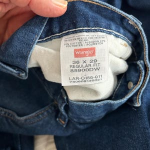 Pu&ograve; includere: Un primo piano dell'interno di un paio di jeans blu denim con un'etichetta Wrangler. L'etichetta mostra la taglia 36 x 29, Regular Fit, 85900DW, LAR-0156-011, 760609135021. L'etichetta indica anche che i jeans sono realizzati in Messico con tessuto proveniente dagli Stati Uniti e che sono composti da 90% cotone e 20% poliestere.