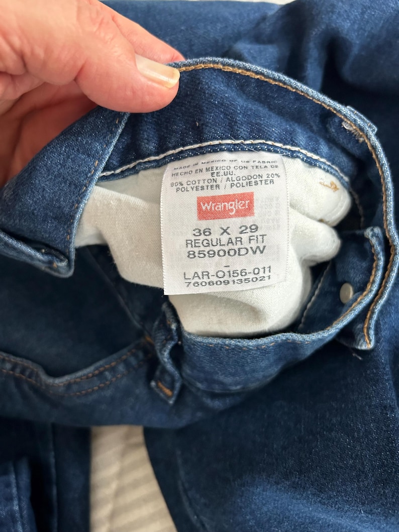 Pu&ograve; includere: Un primo piano dell'interno di un paio di jeans blu denim con un'etichetta Wrangler. L'etichetta mostra la taglia 36 x 29, Regular Fit, 85900DW, LAR-0156-011, 760609135021. L'etichetta indica anche che i jeans sono realizzati in Messico con tessuto proveniente dagli Stati Uniti e che sono composti da 90% cotone e 20% poliestere.