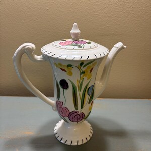 May include: White ceramic teapot with a floral design featuring pink, purple, and yellow flowers. The teapot has a curved handle, a spout, and a lid with a small pink finial. The base has a decorative black trim.