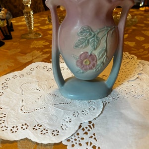 Puede incluir: Un jarrón de cerámica vintage azul y rosa con un diseño floral. El jarrón tiene dos asas y una boca ancha.