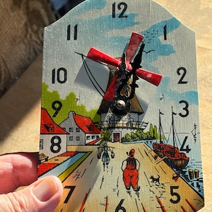 May include: Wooden wall clock with a painted scene of a Dutch village, featuring a windmill with red blades, houses, and figures. The clock face has black numerals and hands. The clock is approximately 8 inches tall.