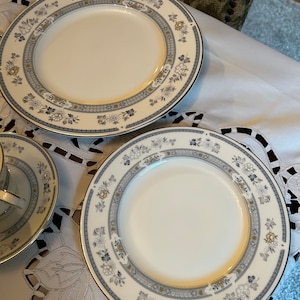 May include: Two white ceramic dinner plates with a blue and gold floral pattern. The plates are on a white tablecloth with a lace overlay.