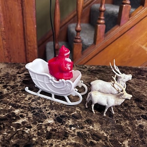 以下が含まれることがあります： 白いそりに乗った赤いサンタクロースのフィギュアと、2つの白いトナカイのフィギュア。そりは曲線的なデザインです。クリスマスのホリデーデコレーション。