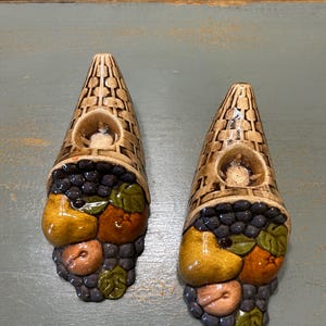 May include: Two ceramic cornucopia-shaped candle holders. The candle holders are brown with a basket weave design. The base of each holder is decorated with colorful fruit, including grapes, pears, and peaches. The candle holders are on a blue-green surface.