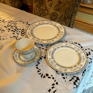 May include: A white tablecloth with a lace pattern, a white teacup and saucer with a blue and gold floral design, and a white dinner plate with a blue and gold floral design.
