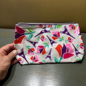 Puede incluir: Bolsa de cosméticos blanca con cierre de cremallera, con un estampado de torres Eiffel moradas, flores rosas y rojas y hojas verdes. La bolsa tiene un acabado brillante y una cremallera morada.