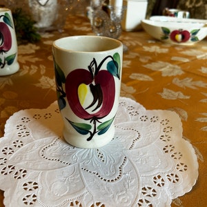 May include: A white ceramic cup with a red apple design on a white doily. The cup has a green stem and leaves on the apple. The doily is on a gold tablecloth.