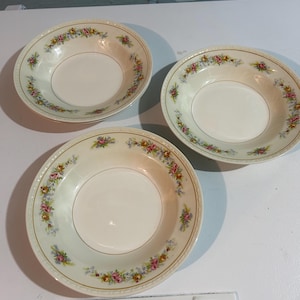 Könnte beinhalten: Drei cremefarbene Keramikschalen mit einem Blumenmuster am Rand. Die Schalen haben einen goldenen Rand und einen weißen Innenraum. Das Blumendesign umfasst rosa, gelbe und blaue Blumen mit grünen Blättern. Die Schalen sind auf einer weißen Oberfläche angeordnet.